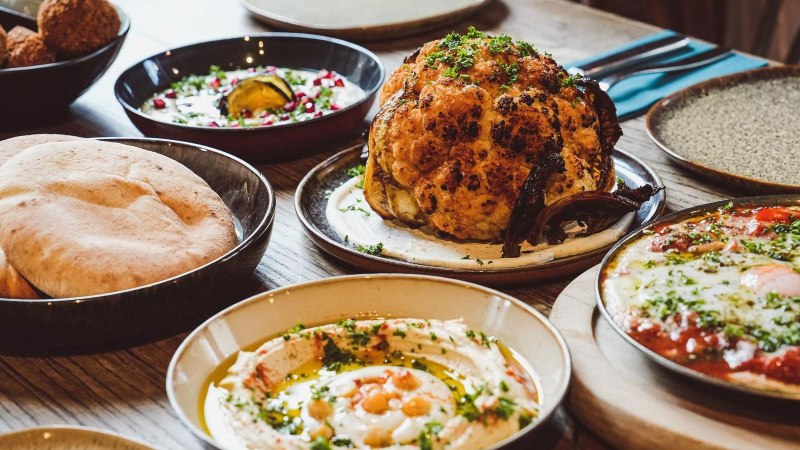 A table set with various Middle Eastern dishes, including roasted cauliflower, hummus with chickpeas, pita bread, and shakshuka with poached eggs in tomato sauce., &copy; yafa, Stuttgart