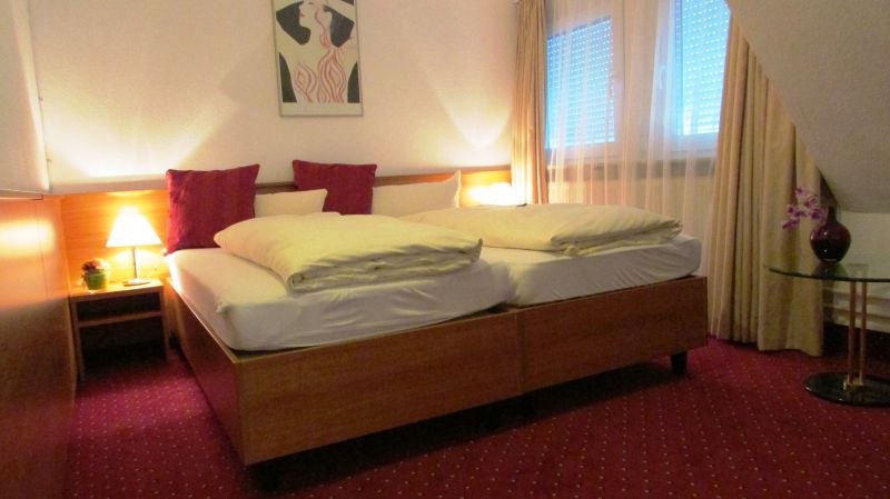 Hotelzimmer mit Doppelbett, rotem Teppich, Nachttischlampe und Bild an der Wand. Fenster mit Vorhängen, Tisch mit Vase., © TOMAS Hotelzimmer mit Doppelbett, rotem Teppich, Nachttischlampe und Bild an der Wand. Fenster mit Vorhängen, Tisch mit Vase., © TOMAS