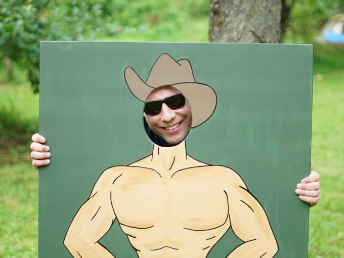 Person behind painted panel with cowboy motif, face visible through hole. Background: green lawn, tree., © Cool-Tours StattReisen Person behind painted panel with cowboy motif, face visible through hole. Background: green lawn, tree., © Cool-Tours StattReisen