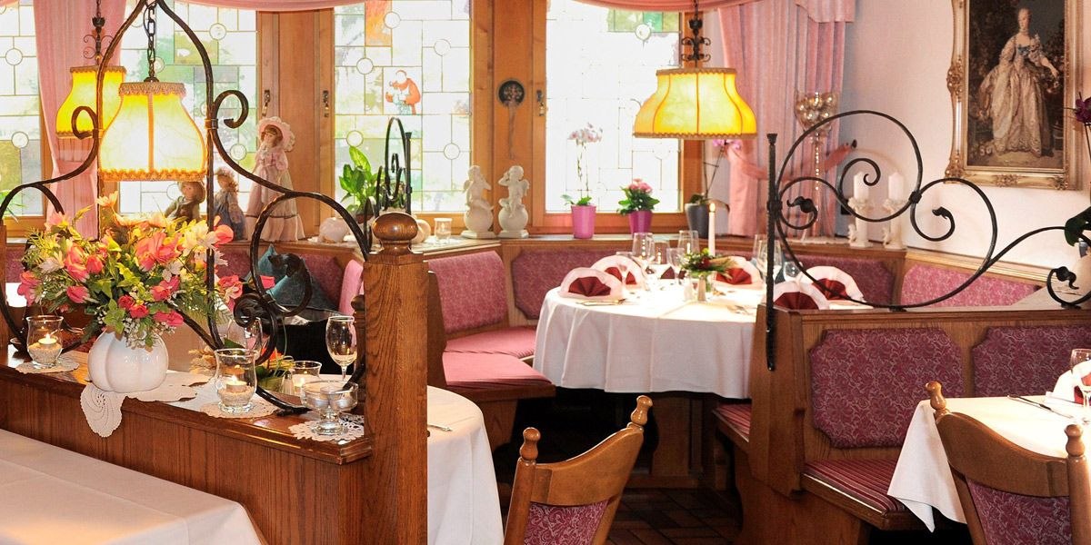 A cozy restaurant with wooden furniture, flower arrangements and decorative lamps. The tables are elegantly laid and there are colorful window decorations., &copy; Hotel Lamm Hegnach