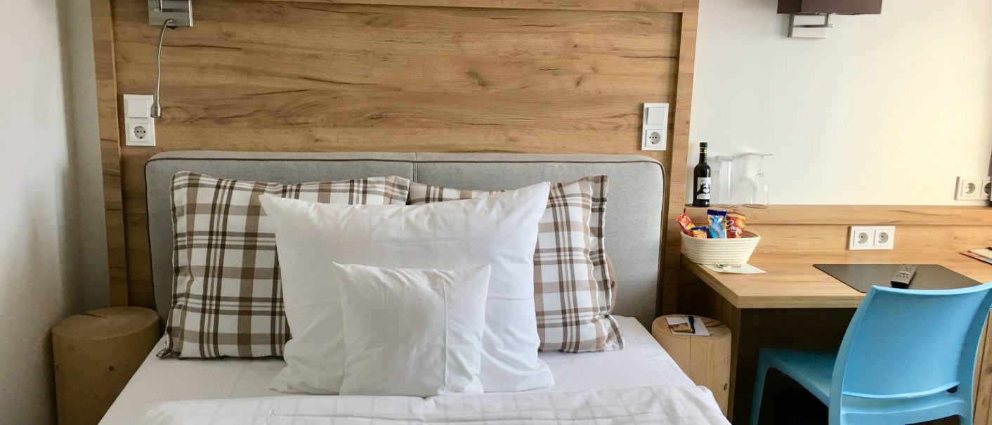 Modern hotel room with single bed, wooden headboard, desk, blue chair and snacks on the table., © TOMAS