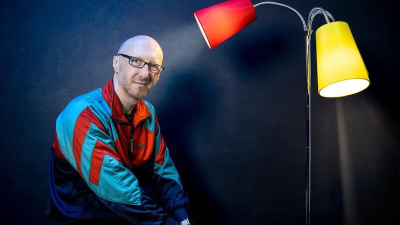A man in a colorful jacket sits next to a floor lamp with red and yellow shades in front of a dark background., &copy; Rosenau Kultur e.V.