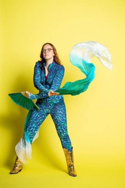 Eine Person in einem schuppenartigen, bunten Kost&uuml;m mit goldenen Stiefeln schwingt bunte F&auml;cher vor einem gelben Hintergrund., &copy; Rosenau Kultur e.V.