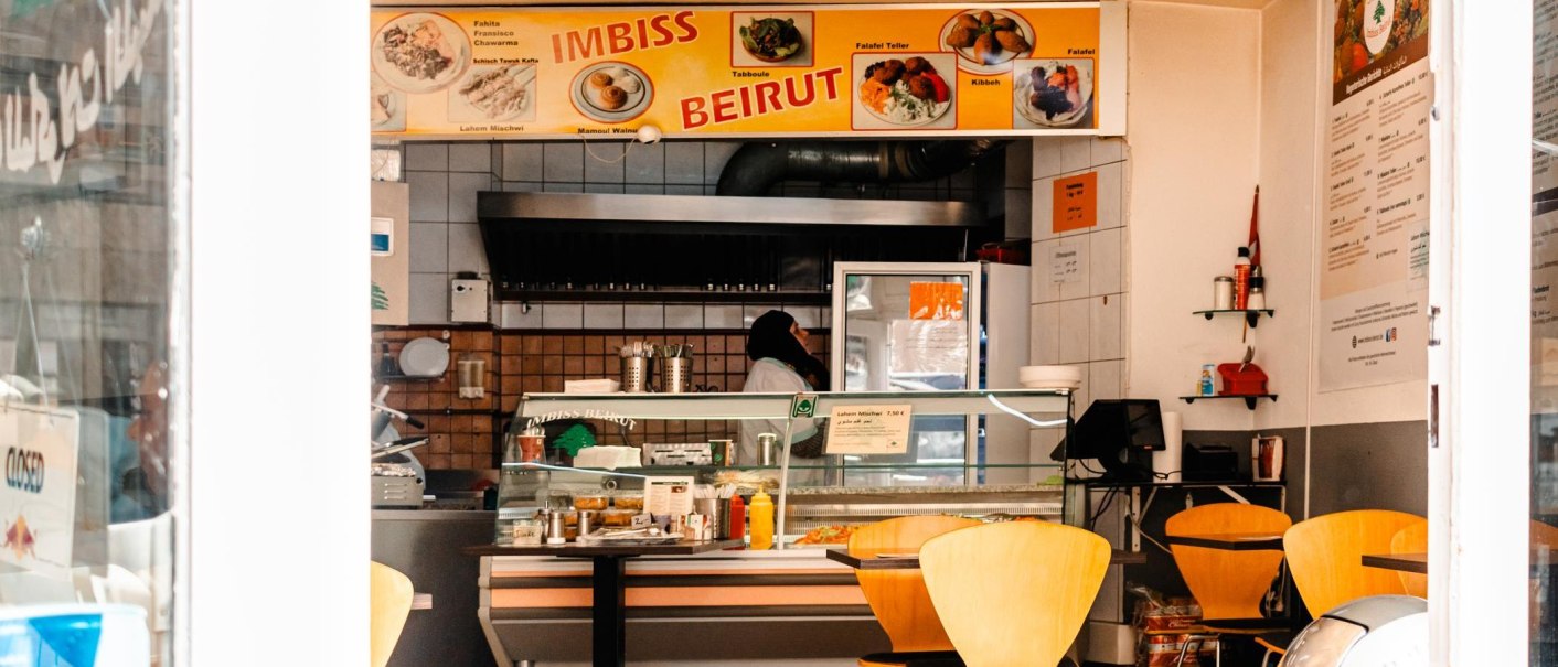 A snack bar with a yellow 'Imbiss Beirut' sign, empty tables and a person behind the counter., © Stuttgart Marketing GmbH, Sarah Schmid A snack bar with a yellow 'Imbiss Beirut' sign, empty tables and a person behind the counter., © Stuttgart Marketing GmbH, Sarah Schmid