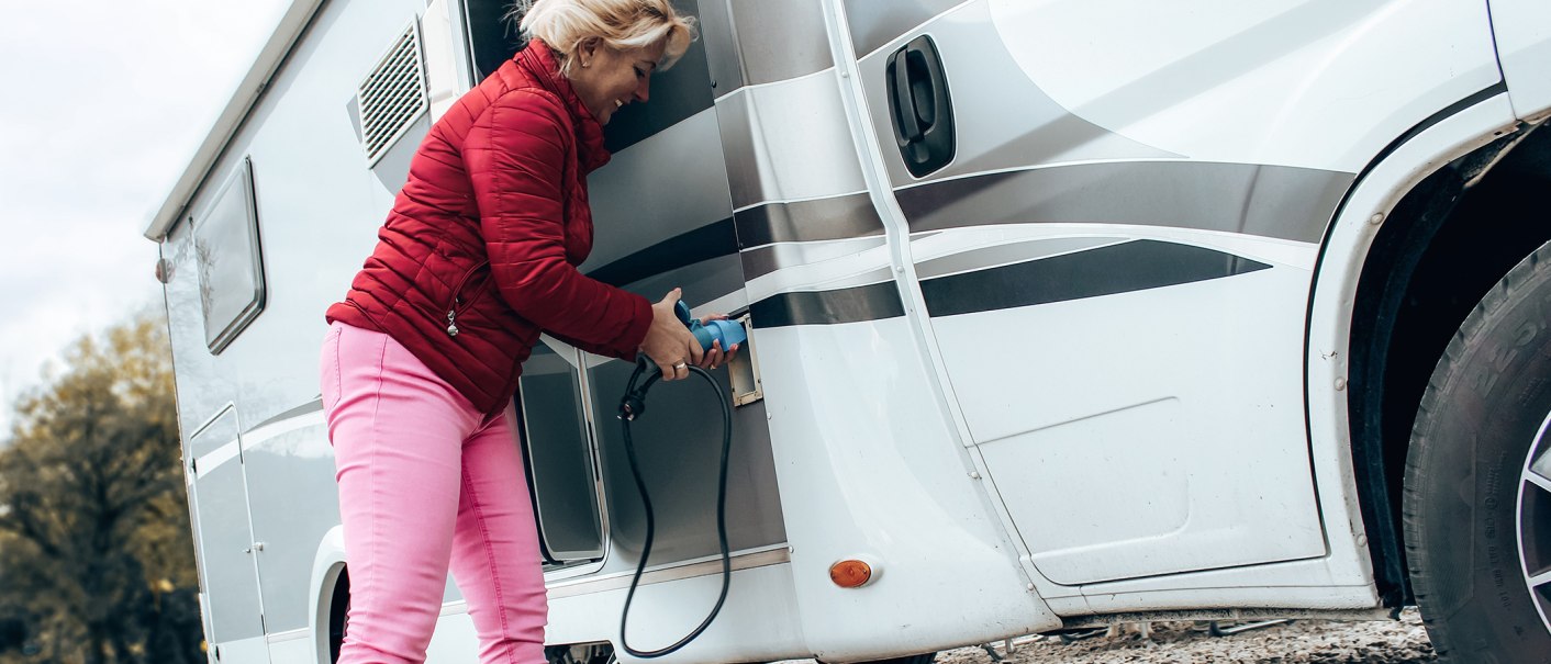 Eine Frau in roter Jacke und pinker Hose schließt ein Wohnmobil an eine Stromquelle an. Sie hält ein blaues Kabel in der Hand., © Erlebnisregion Schwäbischer Albtrauf e. V.