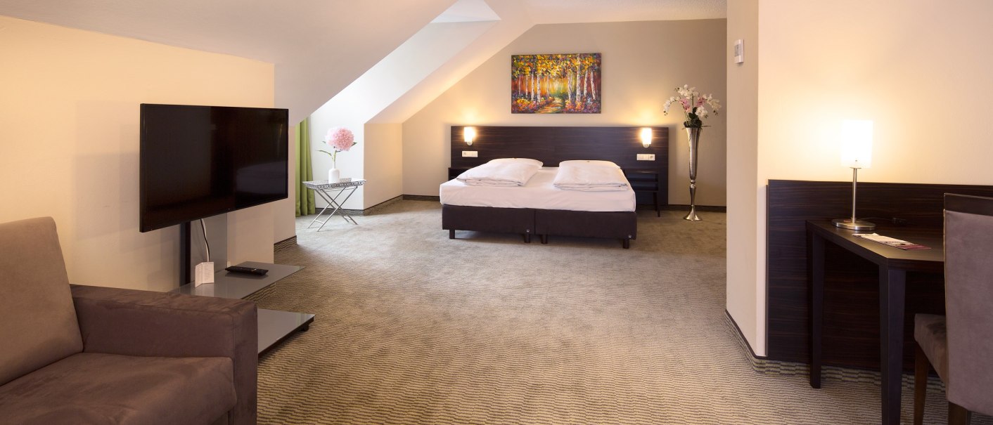 Spacious hotel room with double bed, sofa, flat-screen TV and desk. Light-colored walls, carpeted floor and decorative plants., © Erikson Hotel