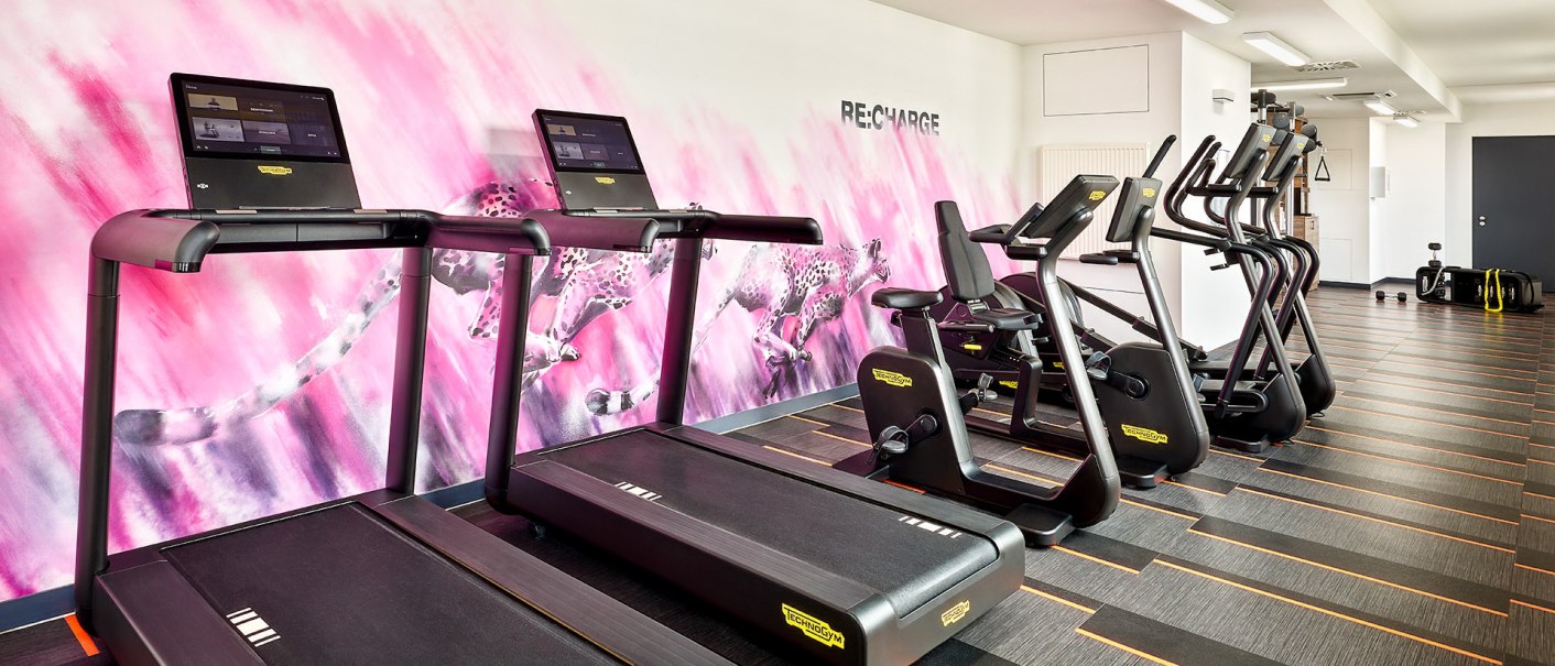 Modern fitness studio with treadmills and ergometers in front of a pink wall with a cheetah motif. The room is bright and inviting., &copy; Aloft Stuttgart