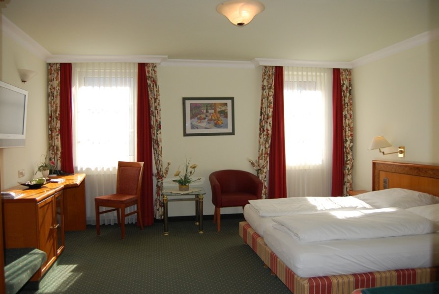 Helles Hotelzimmer mit Doppelbett, Schreibtisch, Stuhl, Sessel und dekorativen Vorhängen. Ein Bild hängt an der Wand., © Jeanette Bender