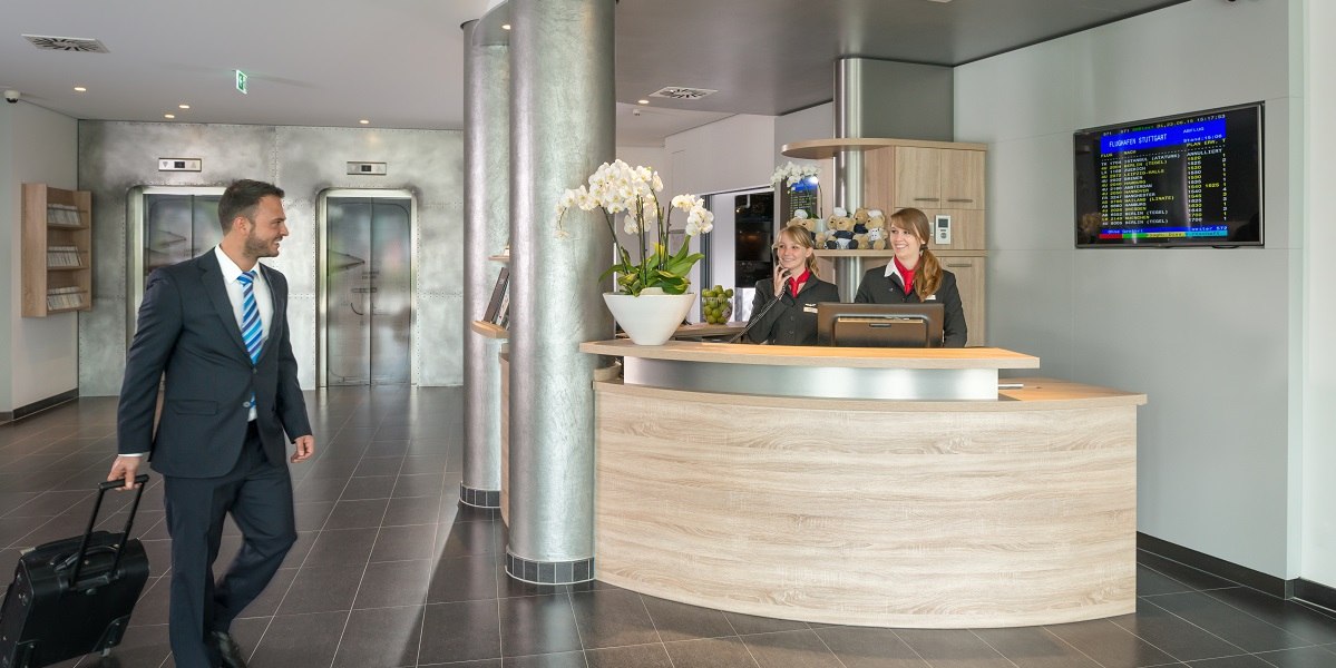 Ein Mann mit Koffer betritt eine moderne Hotel-Lobby. Zwei Rezeptionistinnen stehen hinter einem Empfangstresen. Im Hintergrund sind Aufz&uuml;ge zu sehen., &copy; Essential by Dorint