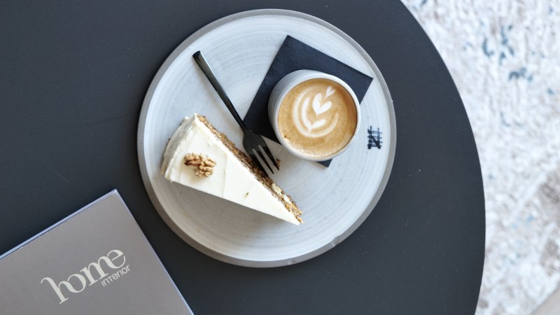 Latte mit Herz-Latte-Art und ein St&uuml;ck Kuchen auf einem Teller auf schwarzem Tisch, daneben eine Karte mit &bdquo;home interior&ldquo;., &copy; Taraba