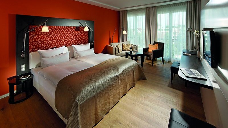 Modernes Hotelzimmer mit großem Bett, oranger Wand, Sofa, Schreibtisch und Fernseher. Helle Vorhänge und Holzboden schaffen eine gemütliche Atmosphäre., © Parkhotel Stuttgart Messe-Airport Modernes Hotelzimmer mit großem Bett, oranger Wand, Sofa, Schreibtisch und Fernseher. Helle Vorhänge und Holzboden schaffen eine gemütliche Atmosphäre., © Parkhotel Stuttgart Messe-Airport