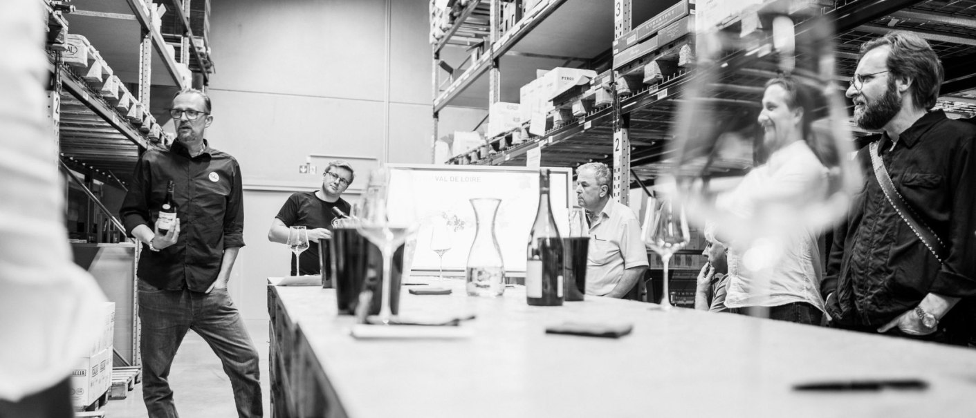 A group of people are standing in a storage room taking part in a wine tasting. Wine glasses and bottles can be seen in the foreground., &copy; club traube