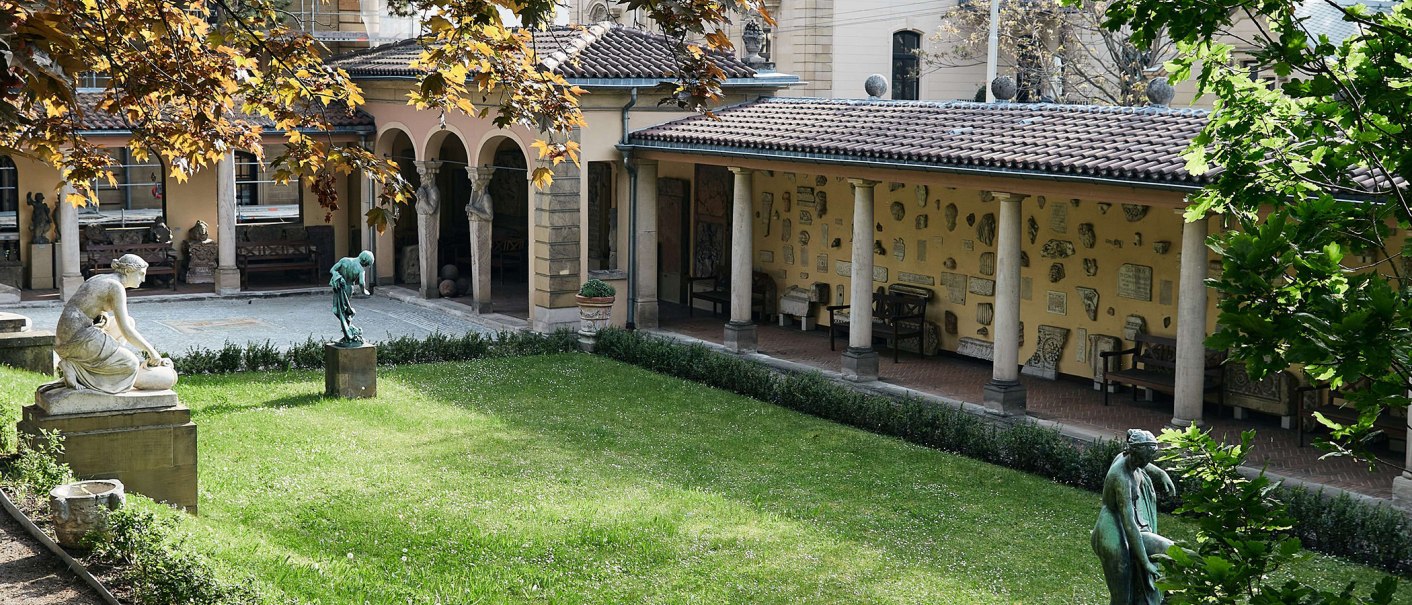 Ein ruhiger Innenhof mit Statuen und antiken Relikten an den Wänden. Der Rasen ist gepflegt, und Bäume spenden Schatten. Arkaden umrahmen den Hof., © Städtisches Lapidarium, Foto Julia Ochs