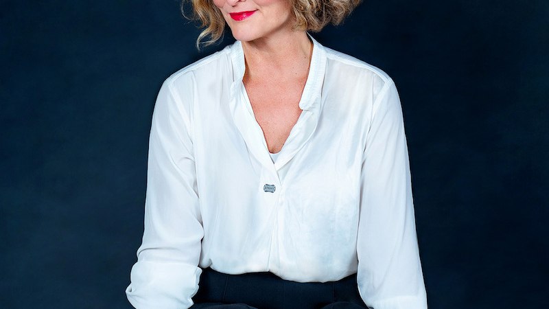 A woman with blonde curly hair wears a white blouse and black trousers. She sits smiling in front of a dark background., &copy; Theaterhaus Stuttgart e.V.