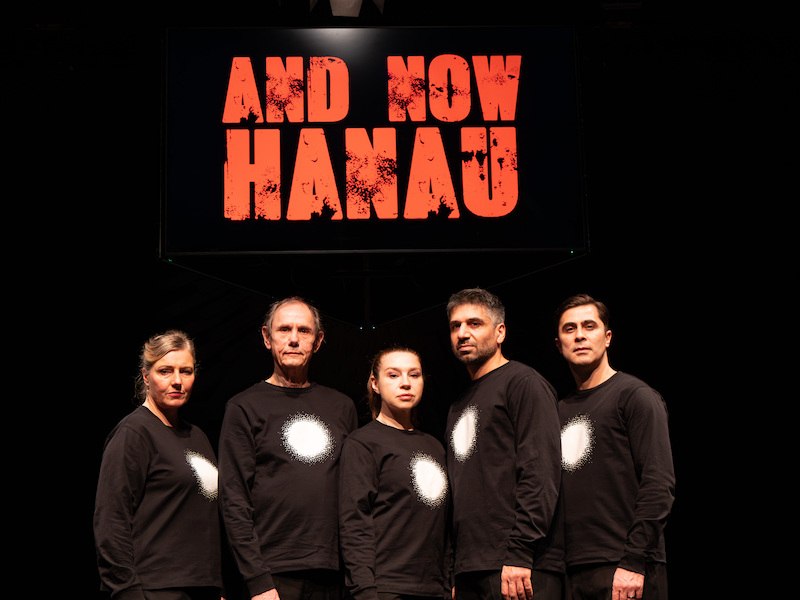 Five people in black shirts with white circles stand in front of a sign that reads 'And Now Hanau'., &copy; Theaterhaus Stuttgart e.V.