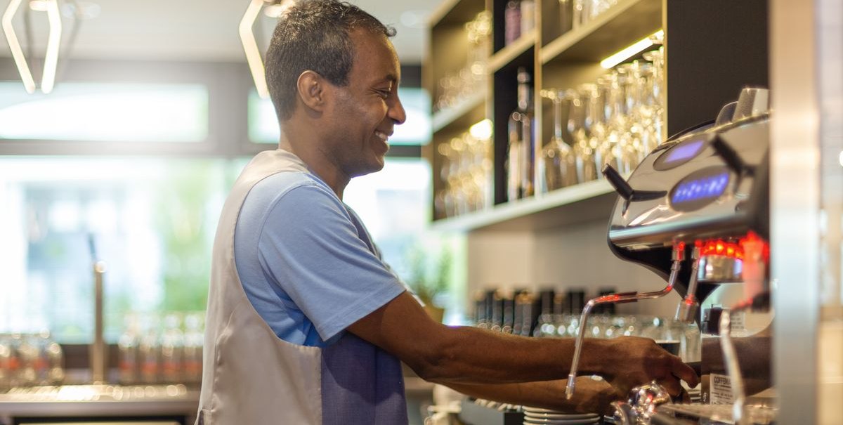 A man is smiling as he works at an espresso machine in a modern bar with glasses in the background., &copy; Novum Hospitality