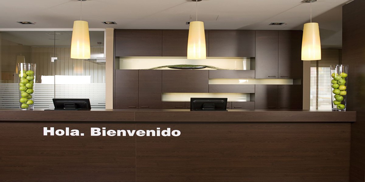 A modern reception area with dark wood furniture, two computers, and the sign "Hola. Bienvenido." Two vases with green apples serve as decoration., © NH Ludwigsburg A modern reception area with dark wood furniture, two computers, and the sign "Hola. Bienvenido." Two vases with green apples serve as decoration., © NH Ludwigsburg
