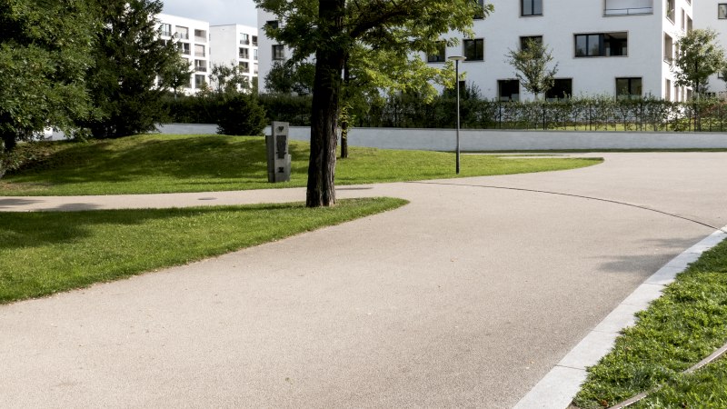 Ein gepflegter Park mit geschwungenen Gehwegen, B&auml;umen und modernen Wohngeb&auml;uden im Hintergrund. Ein Gedenkstein ist sichtbar., &copy; Foto-Kunst Andreas Keller