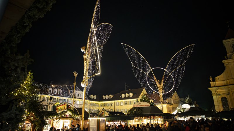 Der Ludwigsburger Barock-Weihnachtsmarkt auf dem Marktplatz bei Nacht, beleuchtete gro&szlig;e Engelsfiguren und Buden. Einige Besucherinnen und Besucher tummeln sich auf dem Platz., &copy; Stuttgart Marketing GmbH, Sarah Schmid