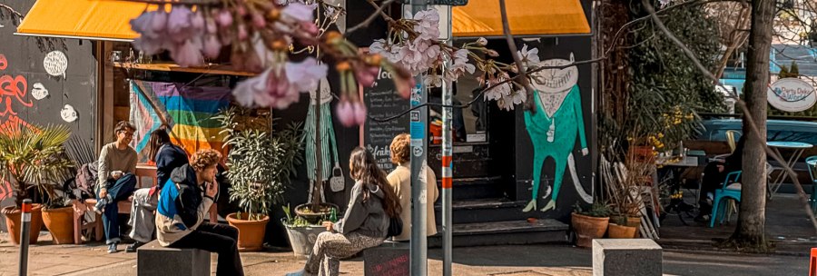 A cozy street caf&eacute; with a yellow sunshade, rainbow flag and blooming cherry blossoms. People sit relaxed in front of it., &copy; Caf&eacute; Moody, Stuttgart