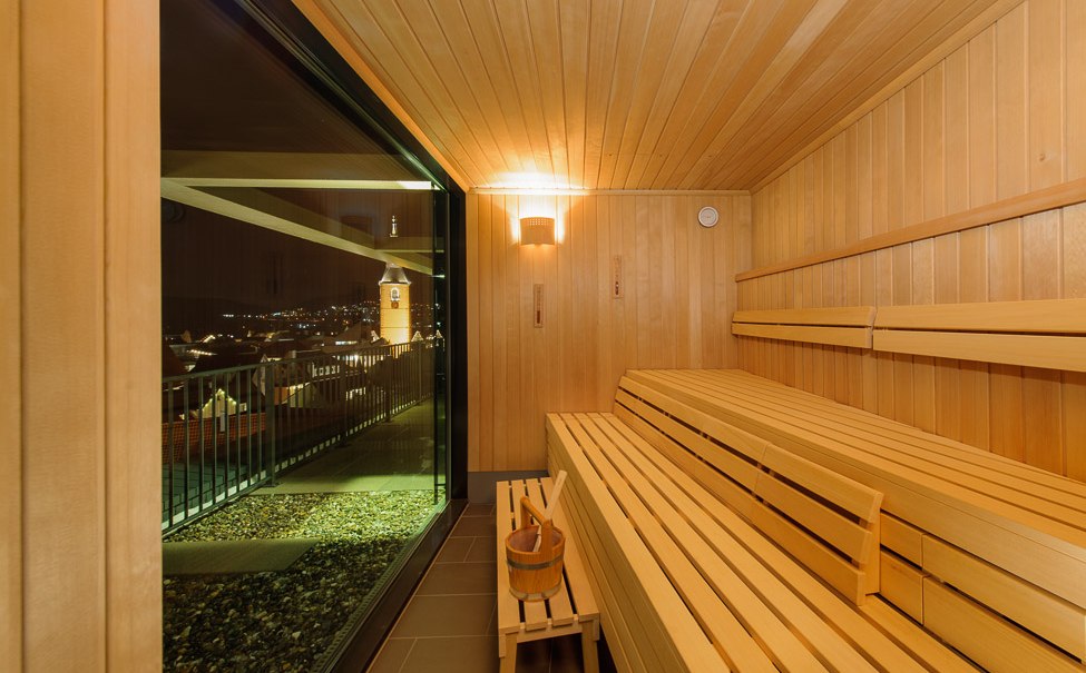 Sauna mit Holzbänken, Eimer und Kelle. Großes Fenster bietet nächtlichen Ausblick auf eine Stadt mit beleuchtetem Turm., © TOMAS Sauna mit Holzbänken, Eimer und Kelle. Großes Fenster bietet nächtlichen Ausblick auf eine Stadt mit beleuchtetem Turm., © TOMAS