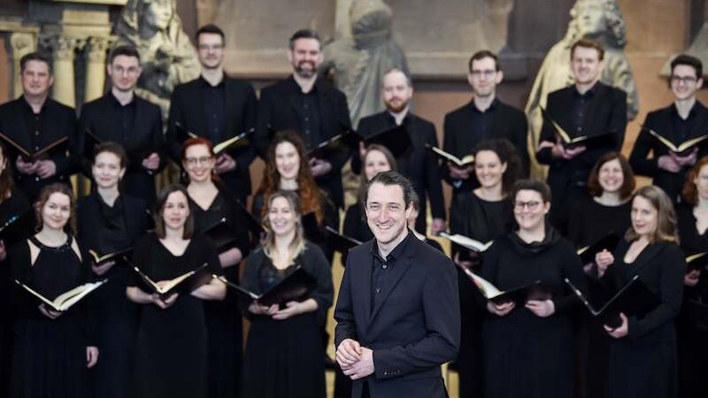 Ein Chor in schwarzer Kleidung singt in einer Kirche. Ein Mann steht lächelnd im Vordergrund. Im Hintergrund sind Statuen und Kirchenfenster zu sehen., © Württembergischer Kammerchor e.V. Ein Chor in schwarzer Kleidung singt in einer Kirche. Ein Mann steht lächelnd im Vordergrund. Im Hintergrund sind Statuen und Kirchenfenster zu sehen., © Württembergischer Kammerchor e.V.