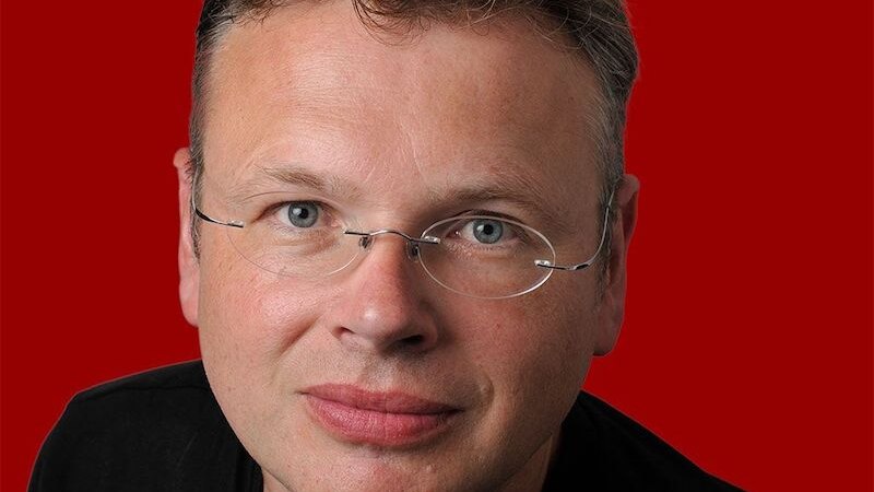 Ein Mann mit Brille und kurzem Haar vor einem roten Hintergrund. Er trägt ein schwarzes Oberteil und blickt direkt in die Kamera., © Börsenverein des Deutschen Buchhandels, Landesverband Baden-Württemberg e.V. Ein Mann mit Brille und kurzem Haar vor einem roten Hintergrund. Er trägt ein schwarzes Oberteil und blickt direkt in die Kamera., © Börsenverein des Deutschen Buchhandels, Landesverband Baden-Württemberg e.V.