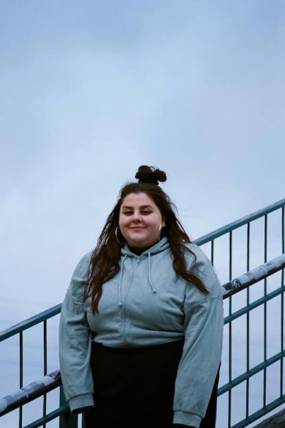 A woman in a blue hooded jacket stands smiling on a staircase. The sky is cloudy and the railing is visible., &copy; Rosenau Kultur e.V.