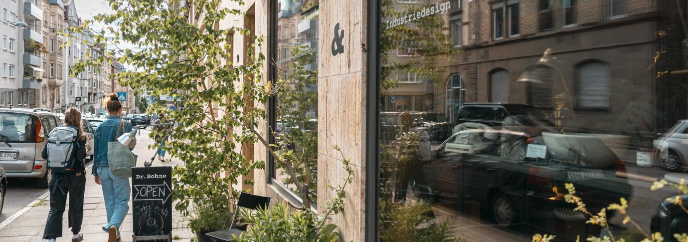 Zwei Personen gehen an einem Gesch&auml;ft mit der Aufschrift 'Dr. Bohne' vorbei. Die Stra&szlig;e ist von Autos ges&auml;umt, und Pflanzen stehen vor dem Schaufenster., &copy; SMG, Sarah Schmid