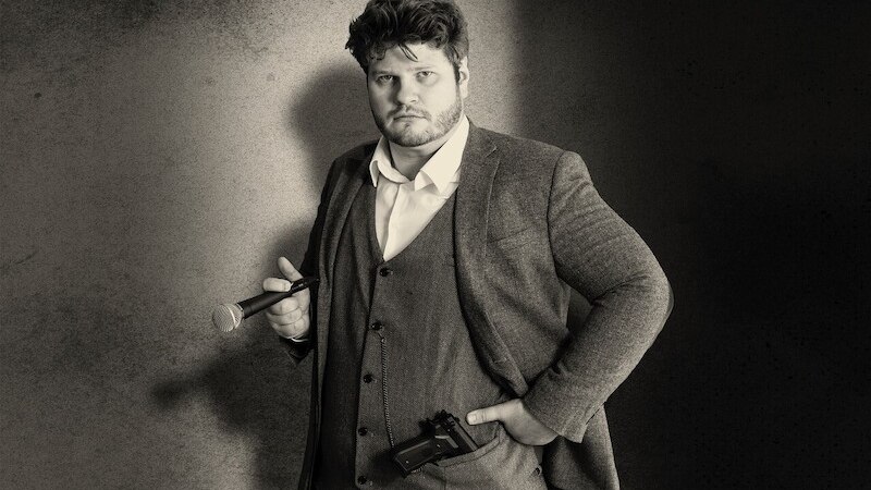 A man in a suit is holding a microphone and a camera. He stands in front of a neutral background and looks seriously into the camera., &copy; Rosenau Kultur e.V.