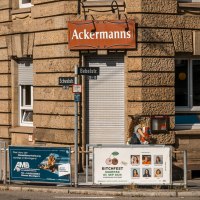 Eckgebäude mit dem Schild 'Ackermanns' an der Fassade. Straßenschilder für Bebelstr. und Schwabstr. sind sichtbar, ebenso mehrere Werbeplakate., © SMG, Sarah Schmid Eckgebäude mit dem Schild 'Ackermanns' an der Fassade. Straßenschilder für Bebelstr. und Schwabstr. sind sichtbar, ebenso mehrere Werbeplakate., © SMG, Sarah Schmid