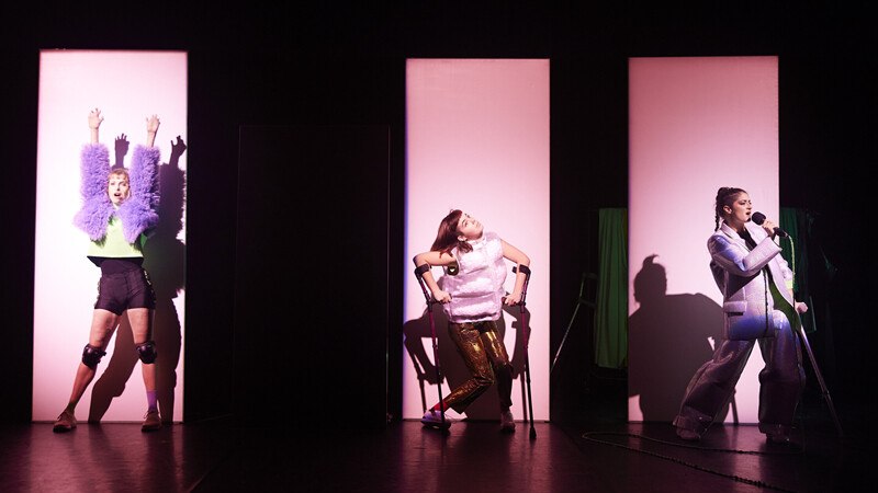 Drei Personen auf einer Bühne in auffälliger Kleidung, beleuchtet von rosa Licht. Eine Person singt in ein Mikrofon, die anderen posieren., © Württembergische Staatstheater Stuttgart