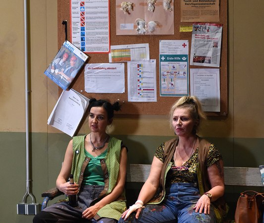 Zwei Frauen in Arbeitskleidung sitzen auf einer Bank vor einer Pinnwand mit Aushängen. Sie wirken nachdenklich und tragen lässige Kleidung., © Theaterhaus Stuttgart e.V. Zwei Frauen in Arbeitskleidung sitzen auf einer Bank vor einer Pinnwand mit Aushängen. Sie wirken nachdenklich und tragen lässige Kleidung., © Theaterhaus Stuttgart e.V.