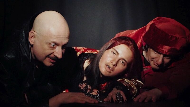 Three people pose in dark surroundings. One is wearing a red robe, one is bald, the third has long hair and dark clothes., &copy; Theater Atelier