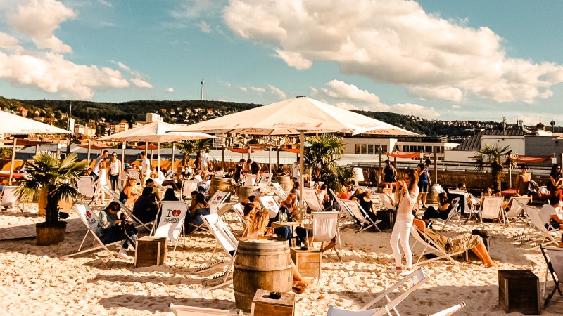 Strandbar mit Liegestühlen und Sonnenschirmen, umgeben von Palmen. Menschen entspannen sich unter blauem Himmel mit Wolken., © Sky Beach