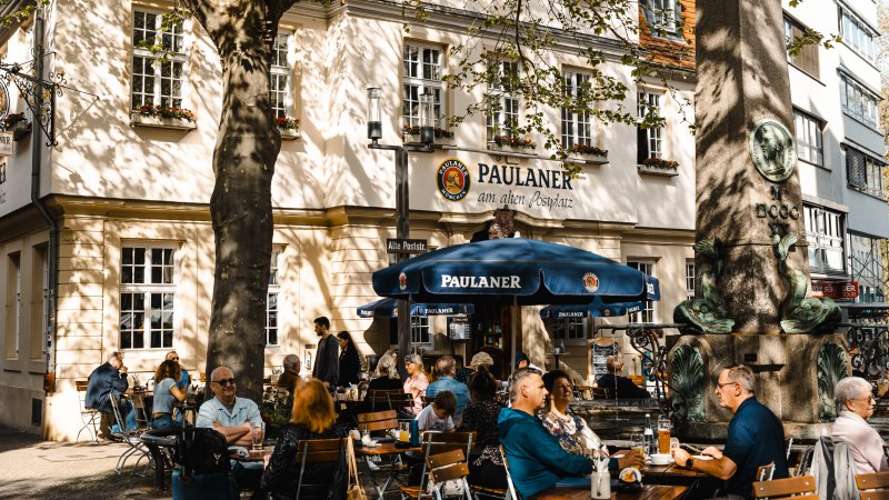 Menschen sitzen im Außenbereich eines Paulaner-Restaurants. Ein großer Baum spendet Schatten, und ein Brunnen ist im Vordergrund zu sehen., © SMG Stuttgart Marketing GmbH - Sarah Schmid Menschen sitzen im Außenbereich eines Paulaner-Restaurants. Ein großer Baum spendet Schatten, und ein Brunnen ist im Vordergrund zu sehen., © SMG Stuttgart Marketing GmbH - Sarah Schmid