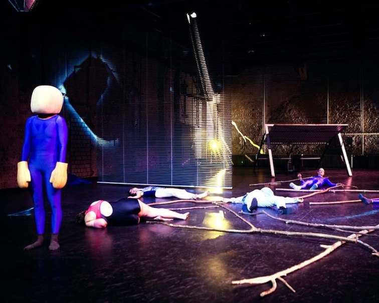 Eine surreale Theaterszene mit einer Figur in einem blauen Anzug und mehreren Personen, die auf dem Boden liegen. Im Hintergrund sind Lichteffekte zu sehen., © Theater Rampe Eine surreale Theaterszene mit einer Figur in einem blauen Anzug und mehreren Personen, die auf dem Boden liegen. Im Hintergrund sind Lichteffekte zu sehen., © Theater Rampe