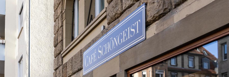 Das Bild zeigt das Schild des Caf&eacute;s Sch&ouml;ngeist an einer steinernen Hausfassade. Im Fenster spiegelt sich ein gegen&uuml;berliegendes Geb&auml;ude., &copy; Stuttgart-Marketing GmbH, Sarah Schmid