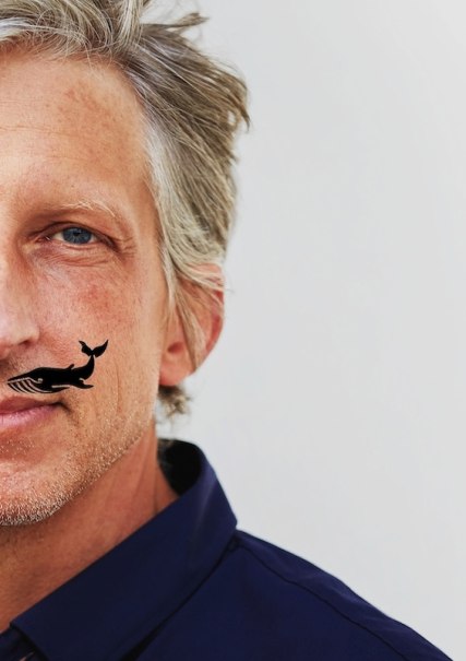 Close-up of a man with gray hair, a blue shirt and a small whale tattoo on his cheek., © Renitenztheater Stuttgart e.V. Close-up of a man with gray hair, a blue shirt and a small whale tattoo on his cheek., © Renitenztheater Stuttgart e.V.