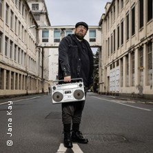 Eine Person steht mit einer Boombox in einer leeren Stra&szlig;e, umgeben von hohen Geb&auml;uden. Die Szene wirkt urban und ruhig., &copy; links im Bild