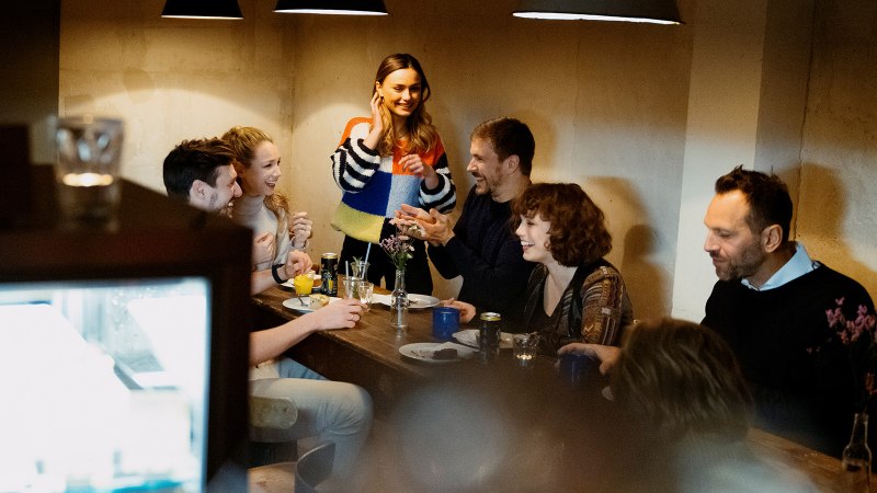 Eine Gruppe von Menschen sitzt in einem Café um einen Tisch und lacht. Eine Frau steht und spricht mit den anderen. Die Atmosphäre ist gemütlich., © Stuttgart Marketing GmbH, Alwin Maigler