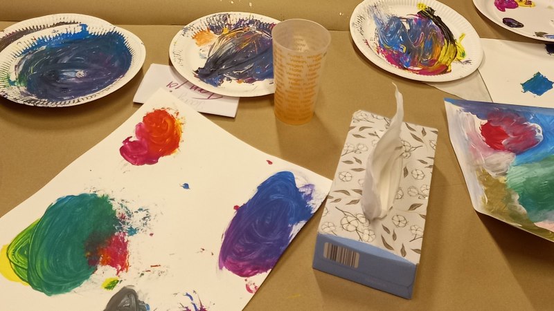 Bunte Farbpaletten und bemalte Blätter liegen auf einem Tisch. Ein Plastikbecher und eine Box mit Papiertüchern sind ebenfalls zu sehen., © KVWH Bunte Farbpaletten und bemalte Blätter liegen auf einem Tisch. Ein Plastikbecher und eine Box mit Papiertüchern sind ebenfalls zu sehen., © KVWH