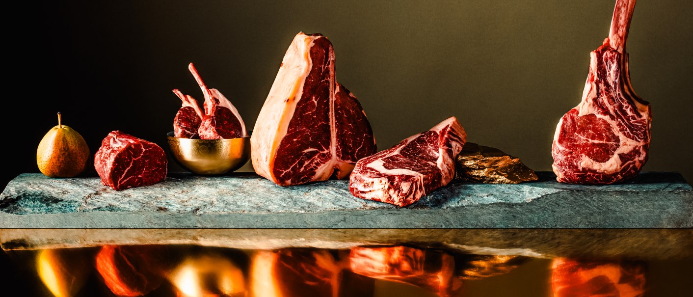 Verschiedene Fleischstücke, darunter Tomahawk-Steaks und Lammkoteletts, sind kunstvoll auf einer Steinplatte arrangiert. Eine Birne liegt daneben., © The Meat Club, Stuttgart