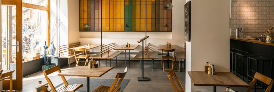 Modernes Café mit Holzmöbeln, buntem Glasfenster und einladender Atmosphäre. Große Fenster lassen viel Licht herein., © SMG, Sarah Schmid Modernes Café mit Holzmöbeln, buntem Glasfenster und einladender Atmosphäre. Große Fenster lassen viel Licht herein., © SMG, Sarah Schmid