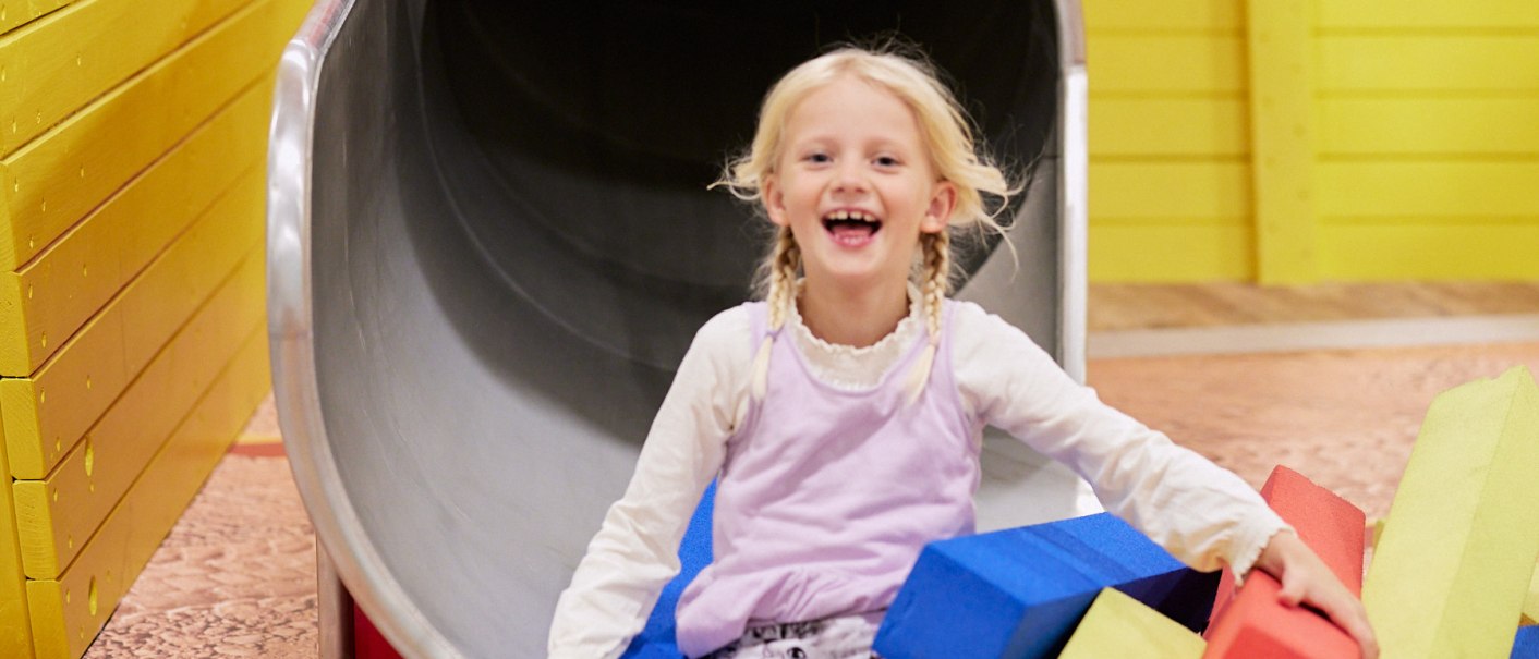 Ein Kind rutscht lachend aus einer orangefarbenen R&ouml;hre auf einer Kinderbaustelle. Umgeben von bunten Schaumstoffbl&ouml;cken in Gelb, Blau und Rot., &copy; Julia Ochs