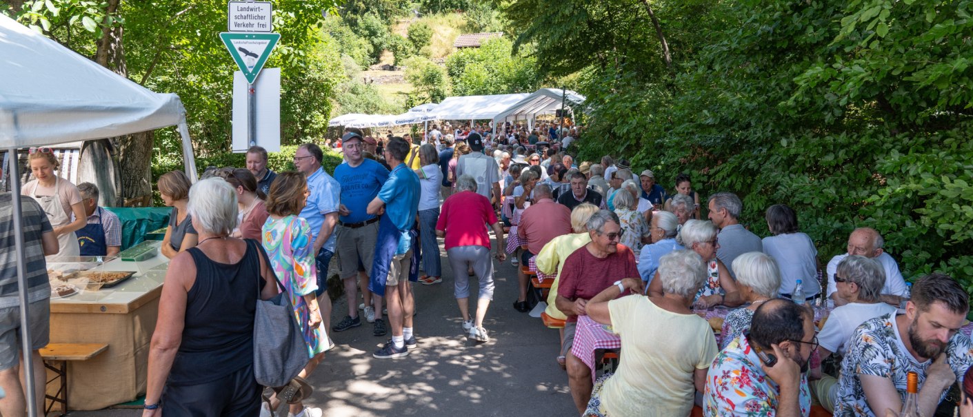 Menschen sitzen und stehen bei einem Fest im Freien. Tische und Zelte sind aufgestellt, umgeben von B&auml;umen. Ein Schild weist auf landwirtschaftlichen Verkehr hin., &copy; Frank Nonnenmann