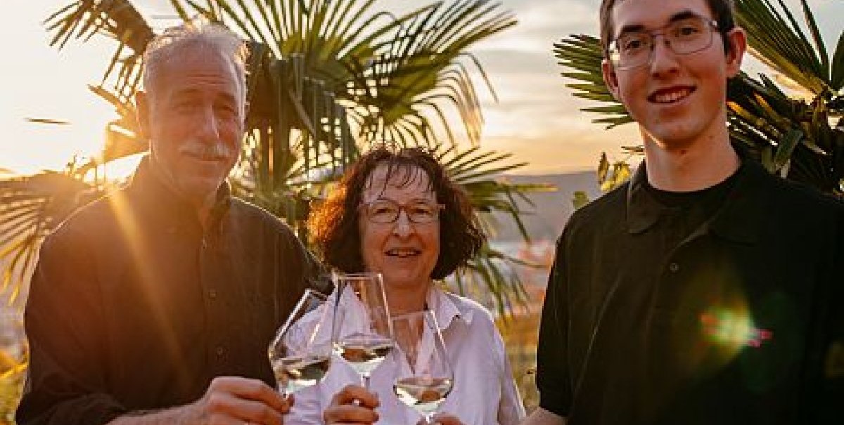 Drei Personen sto&szlig;en mit Weingl&auml;sern an, umgeben von Palmen im Sonnenuntergang., &copy; Weingut Bayer