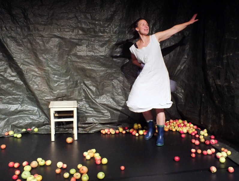 Eine Frau in einem wei&szlig;en Kleid und blauen Gummistiefeln tanzt auf einer B&uuml;hne, die mit &Auml;pfeln bedeckt ist. Ein Hocker steht im Hintergrund., &copy; Dieter Nelle