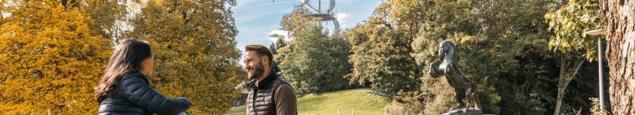Zwei Personen sitzen im H&ouml;henpark Killesberg, umgeben von Herbstb&auml;umen. Im Hintergrund ist der Killesbergturm zu sehen, neben einer Pferdestatue., &copy; Stuttgart-Marketing GmbH, Sarah Schmid