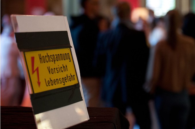 Schild mit Warnung vor Hochspannung, im Vordergrund. Unscharfe Menschenmenge im Hintergrund in einem Innenraum., © Landesmuseum Württemberg